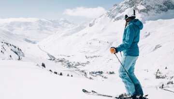 A female skier in blue ski clothing stands on the slope and looks into the distance. | © Daniel Zangerl, Lech Zürs Tourismus