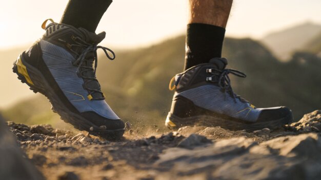 Nahaufnahme von Wanderschuhen auf felsigem Bergpfad bei Sonnenaufgang, Beine einer Person sichtbar, Staub wirbelt auf.