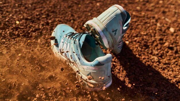 Hellblaue Wanderschuhe stehen auf rotem, erdigem Untergrund, Staub und kleine Steine fliegen herum.