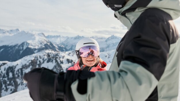 Two skiers on a snowy mountain; in the foreground, a person in a green jacket; in the background, a smiling woman with a white helmet and pink ski goggles against an alpine mountain backdrop. | © INTERSPORT International Corporation GmbH