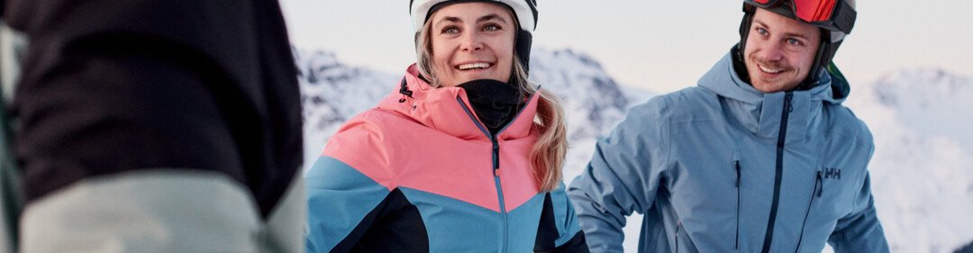 Drei Skifahrer in Helmen und Winterjacken auf einer verschneiten Bergpiste, eine Frau in pink-blauer Jacke lächelt im Vordergrund, ein Mann in blauer Jacke steht rechts und schaut sie an. | © INTERSPORT International Corporation GmbH
