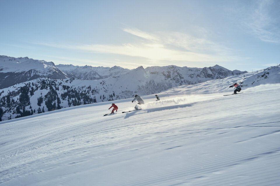 Quatre skieurs en tenue de sports d'hiver descendent en file indienne une piste ensoleillée fraîchement damée, avec les montagnes enneigées des Alpes en arrière-plan. | © INTERSPORT International Corporation GmbH