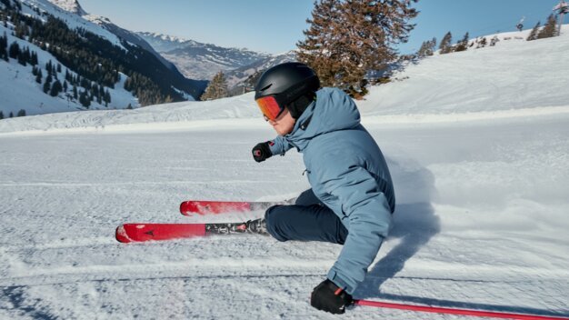 Skieur vêtu d'un anorak bleu et d'un casque noir, équipé de skis rouges, effectuant un virage carvé sur une piste damée devant des montagnes enneigées. | © INTERSPORT International Corporation GmbH