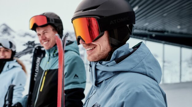 Trois skieurs vêtus de vestes d'hiver et de casques ; au premier plan, un homme souriant avec un casque noir et des lunettes de ski rouges tient des skis rouges, avec en arrière-plan un paysage montagneux enneigé. | © INTERSPORT International Corporation GmbH