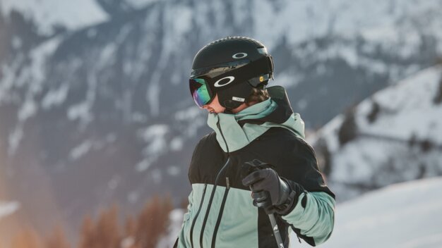 Lunettes de ski OTG Skieur devant un autre skieur portant un casque noir, des lunettes de ski Oakley miroir et une veste noir menthe, tenant un bâton de ski avec des gants devant un paysage montagneux enneigé. | © INTERSPORT International Corporation GmbH