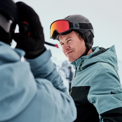 Two people skiing: close-up of a smiling man in a blue-green ski jacket with a helmet and orange ski goggles, the shoulder of a second person in the foreground, snow-covered mountain landscape in the background. | © INTERSPORT International Corporation GmbH