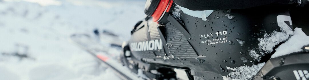 Close-up of a black Salomon ski boot (Flex 110) with snow on a ski binding, person in black ski pants, snowy mountain landscape in the background. | © INTERSPORT International Corporation GmbH