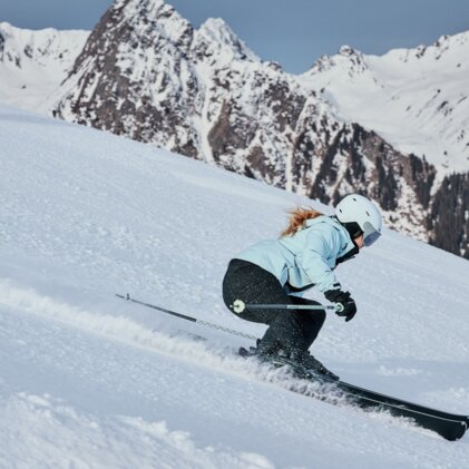 Femme en train de skier, vêtue d'une veste bleu clair et d'un casque blanc, elle descend une piste enneigée escarpée devant des montagnes enneigées. | © INTERSPORT International Corporation GmbH