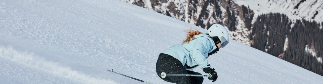 Woman skiing in a light blue jacket and white helmet, she is skiing down a steep, snow-covered slope in front of snow-covered mountains. | © INTERSPORT International Corporation GmbH