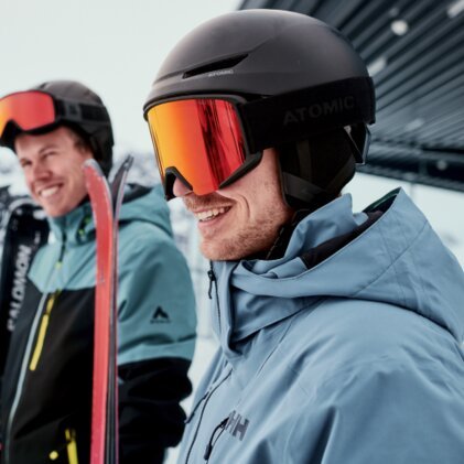 Three skiers in winter jackets and helmets; in the foreground, a smiling man wearing a black helmet and red ski goggles holds red skis, with a snowy mountain backdrop in the background. | © INTERSPORT International Corporation GmbH