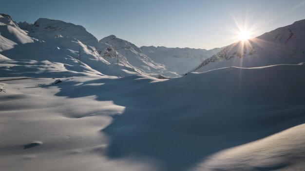 Snow-covered alpine valley at sunrise with sunlit mountain peaks, gentle snow-covered hills, and a trail of footprints in the foreground. | © INTERSPORT International Corporation GmbH