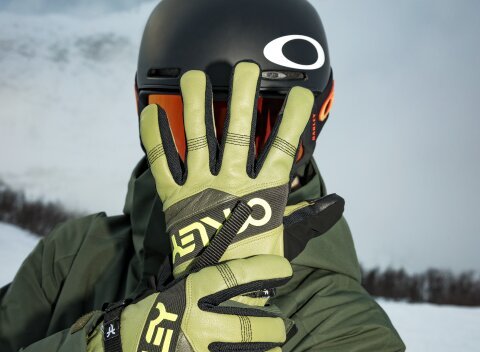 Man in een groene skikleding met bijpassende Oakley-skibril en Oakley-skihandschoenen houdt zijn linkerhand open voor zijn gezicht, terwijl zijn rechterhand zijn linkerpols omvat. | © 30.09.2026