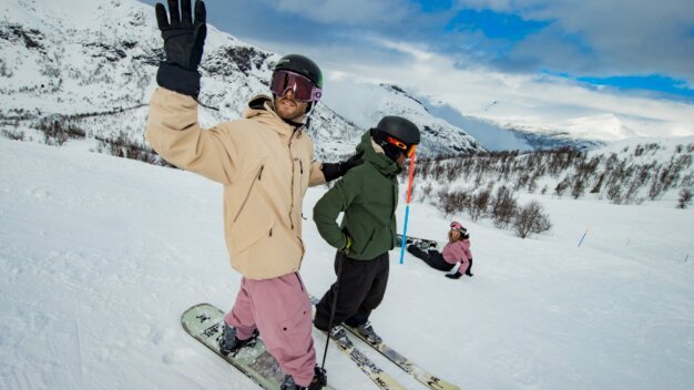 Een man op een snowboard staat naast een skiër op de piste en steekt zijn hand op om te zwaaien.