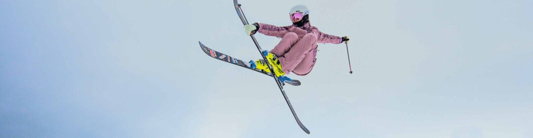 Skifahrende in rosa Skianzug mit Helm und rosa Skibrille führt bei einem Luftsprung gekreuzte Skier vor vor hellem Winterhimmel. | © INTERSPORT International Corporation GmbH