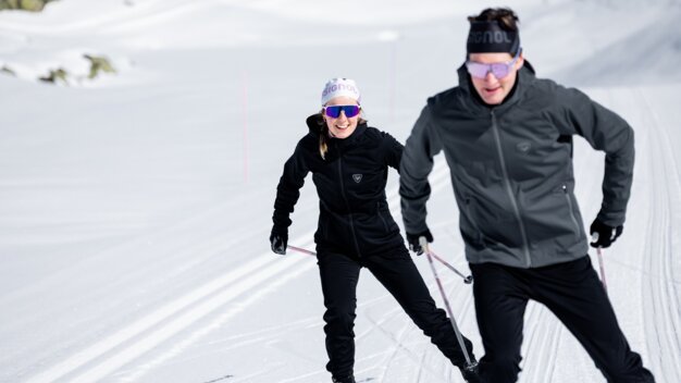 Deux skieurs de fond vêtus de tenues outdoor noires et portant des lunettes de protection, skiant l'un derrière l'autre sur une piste damée ensoleillée. La femme sourit. | © INTERSPORT International Corporation GmbH