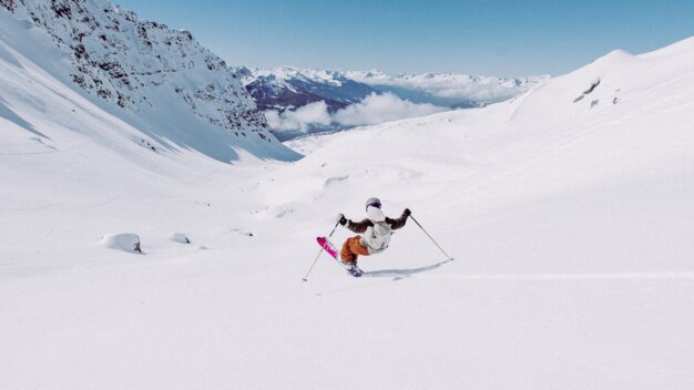 Skiër in witte jas en oranje broek met roze ski's rijdt in diepe sneeuw voor een ver, besneeuwd bergdal. | © INTERSPORT International Corporation GmbH