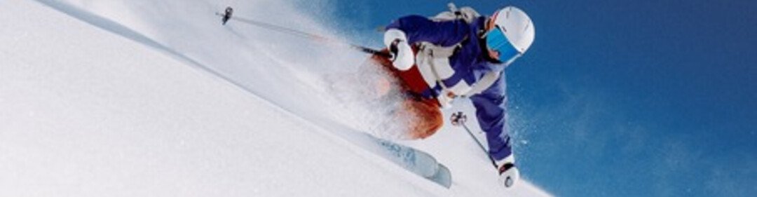 Un skieur vêtu d'une veste bleue et d'un casque blanc descend en diagonale une piste raide recouverte de poudreuse, soulevant des nuages de neige. | © INTERSPORT International Corporation GmbH