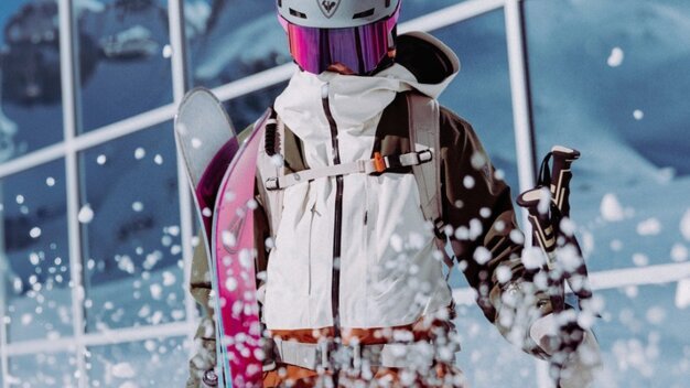 Skieuse vêtue d'une veste blanche, d'un casque et de lunettes de ski violettes, équipée de skis et de bâtons roses ; projections de neige devant une façade vitrée avec reflet des montagnes. | © INTERSPORT International Corporation GmbH