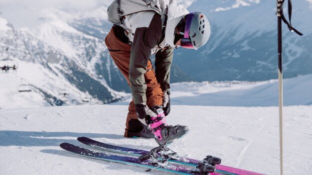 Personne portant un casque de ski et un sac à dos, fixant une boucle de chaussure de ski rose et noire à une fixation sur une piste de montagne enneigée ; regardant vers le bas, montagnes en arrière-plan. | © INTERSPORT International Corporation GmbH