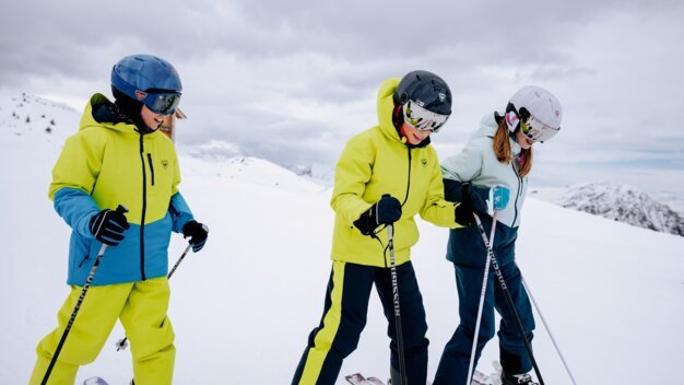 Trois enfants portant des casques et des vestes de ski colorées se tiennent côte à côte, tout sourire, sur leurs skis, sur une piste enneigée. | © INTERSPORT International Corporation GmbH