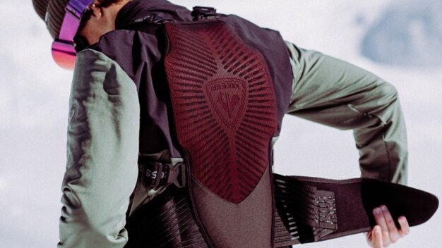 Personne vue de dos, enfilant une protection dorsale rembourrée/un gilet airbag par-dessus un pantalon de ski orange ; elle porte une veste verte, un bonnet marron et des lunettes de ski violettes devant un paysage montagneux enneigé. | © INTERSPORT International Corporation GmbH