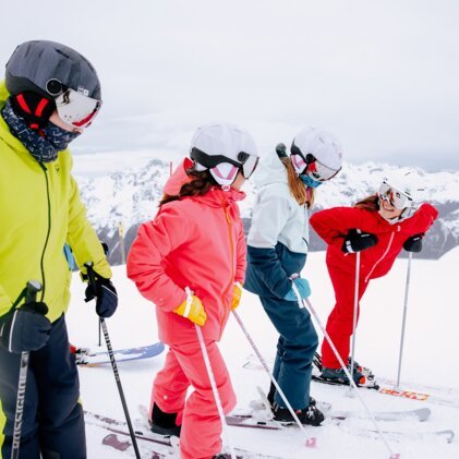 Groupe de quatre skieurs vêtus de vestes et de casques colorés, debout avec leurs bâtons de ski au bord d'une piste enneigée, avec des montagnes enneigées en arrière-plan. | © INTERSPORT International Corporation GmbH