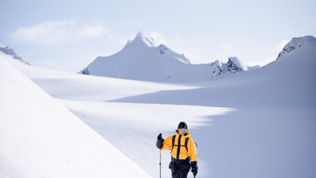Skiër in gele jas met skistokken in een weids, besneeuwd berglandschap voor besneeuwde bergtoppen. | © INTERSPORT International Corporation GmbH