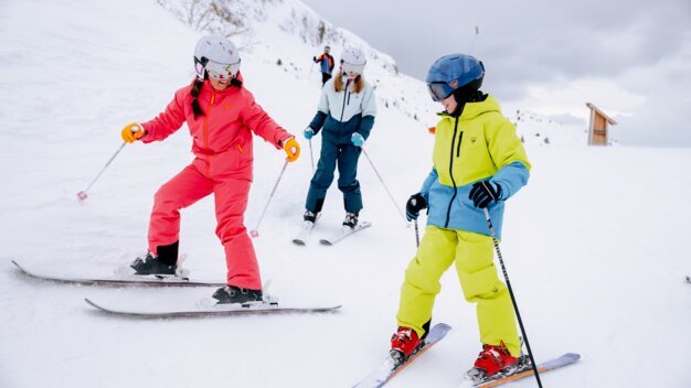 Trois enfants vêtus de combinaisons de ski colorées (rouge, jaune, bleu), équipés de casques et de bâtons de ski, skient sur une piste enneigée avec des montagnes en arrière-plan. | © INTERSPORT International Corporation GmbH