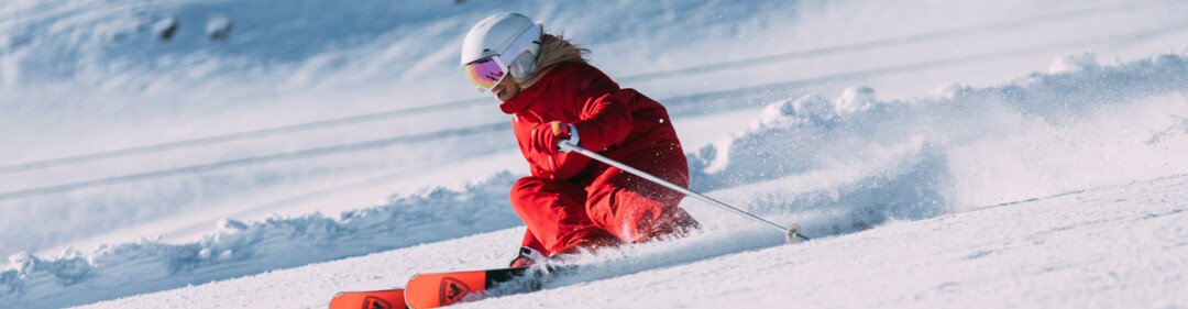 Person in rotem Skianzug und weißem Helm beim Carven auf sonniger, frisch präparierter Skipiste vor schneebedecktem Berg. | © INTERSPORT International Corporation GmbH
