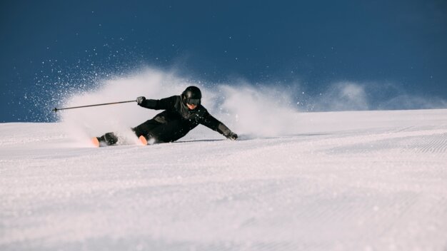 Skieur en tenue noire et casque avec des skis orange effectuant un virage carvé, projections de neige sur une piste damée sous un ciel bleu clair. | © INTERSPORT International Corporation GmbH