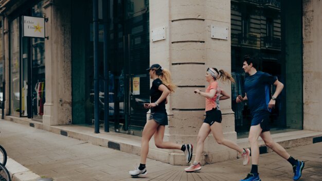 Trois coureurs en tenue de sport (une femme en noir, une femme en rose, un homme en bleu) courent le long d'un coin de rue dans une rue commerçante urbaine. | © INTERSPORT International Corporation GmbH