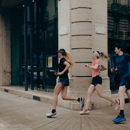 Three runners in sportswear (one in black, one in pink, one man in blue) run along the corner of a building on an urban shopping street. | © INTERSPORT International Corporation GmbH
