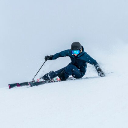 Skier in a dark blue jacket and black helmet with blue ski goggles, carving a deep turn in powder snow and kicking up snow. | © INTERSPORT International Corporation GmbH