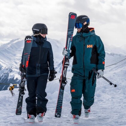 Two skiers wearing helmets and ski goggles walk side by side through deep snow, each carrying skis and wearing winter clothing (HH logo visible); snow-covered mountain landscape in the background. | © INTERSPORT International Corporation GmbH