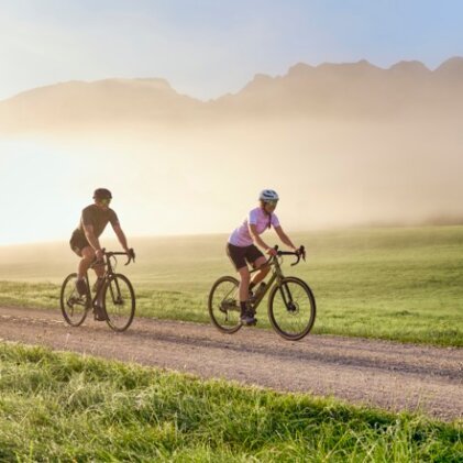 Two cyclists riding on a gravel road through misty scenery, surrounded by soft morning light.