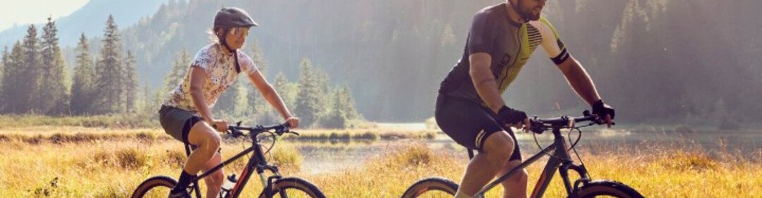 Deux cyclistes roulent dans une prairie à la lumière du soir, entourés de montagnes et de forêt.