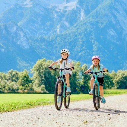 Deux enfants à vélo sur un chemin de gravier, dans un paysage ensoleillé avec des montagnes en arrière-plan.