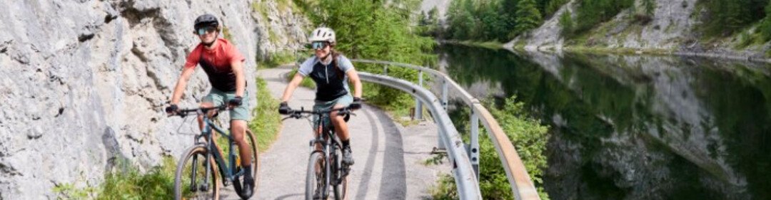 Deux cyclistes sur un chemin étroit le long d’une rivière, entourés de falaises et de nature verdoyante.