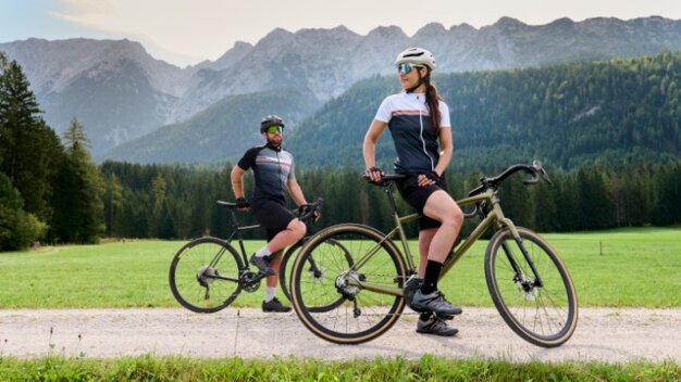 Twee fietsers op een grindpad, omringd door groene velden en een indrukwekkend berglandschap.