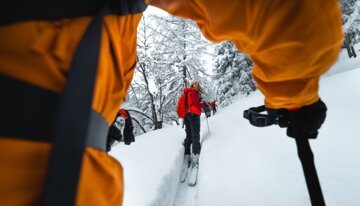 Ski touring in snow, view through a person's arms at a woman in a red jacket. | © Atomic Austria GmbH