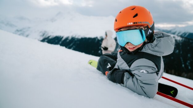 Enfant avec un casque de ski orange et des lunettes bleues assis dans la neige.