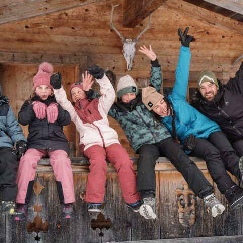 Group of people in winter clothes sitting in front of a wooden cabin and waving. | © INTERSPORT International Corporation GmbH