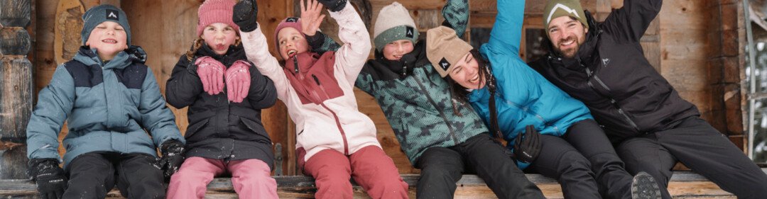 Groep mensen in winterkleding zit voor een houten hut en zwaait. | © INTERSPORT International Corporation GmbH