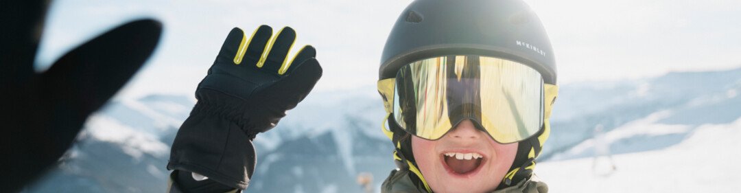 © INTERSPORT International Corporation GmbH Jongen met skibril en helm zwaait op een skipiste. | © INTERSPORT International Corporation GmbH