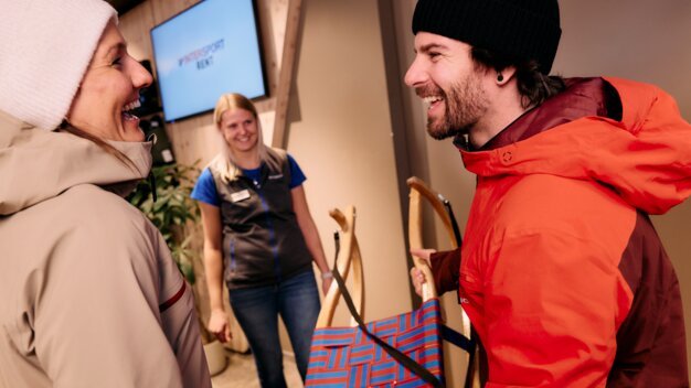 Personnes souriantes avec une luge dans un magasin de location de ski. | © ARMIN WALCHER