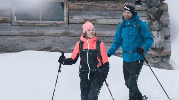 Ein Paar wandert im Schnee vor einem Holzhaus. | © IIC/McKINLEY