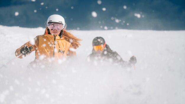 INTERSPORT Rent Helly Hansen Powder Skiing Couple Two skiers in deep snow, the woman in the foreground is wearing a yellow jacket.