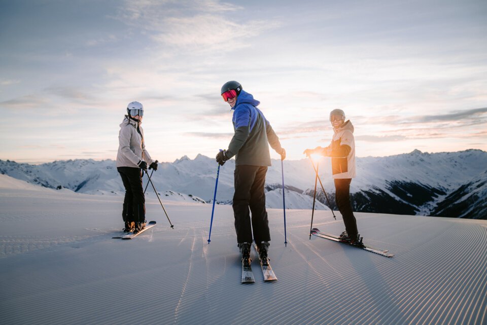 INTERSPORT Rent Salomon Skiing Perfect Ski Day Sunrise Drei Skifahrer auf einer Skipiste vor schneebedeckten Bergen im Sonnenuntergang.