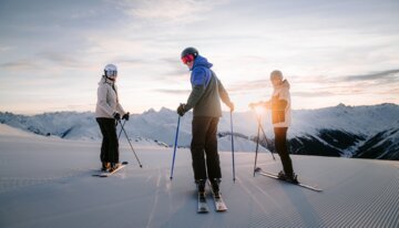 Three skiers on a groomed slope at sunset.