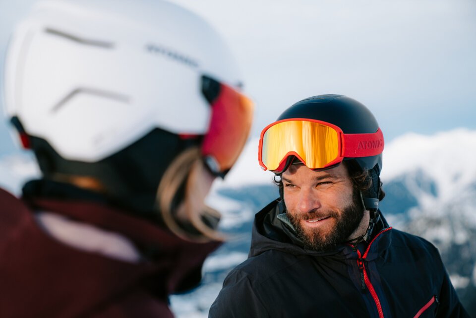 Man with beard and ski goggles smiles at a person wearing a ski helmet.